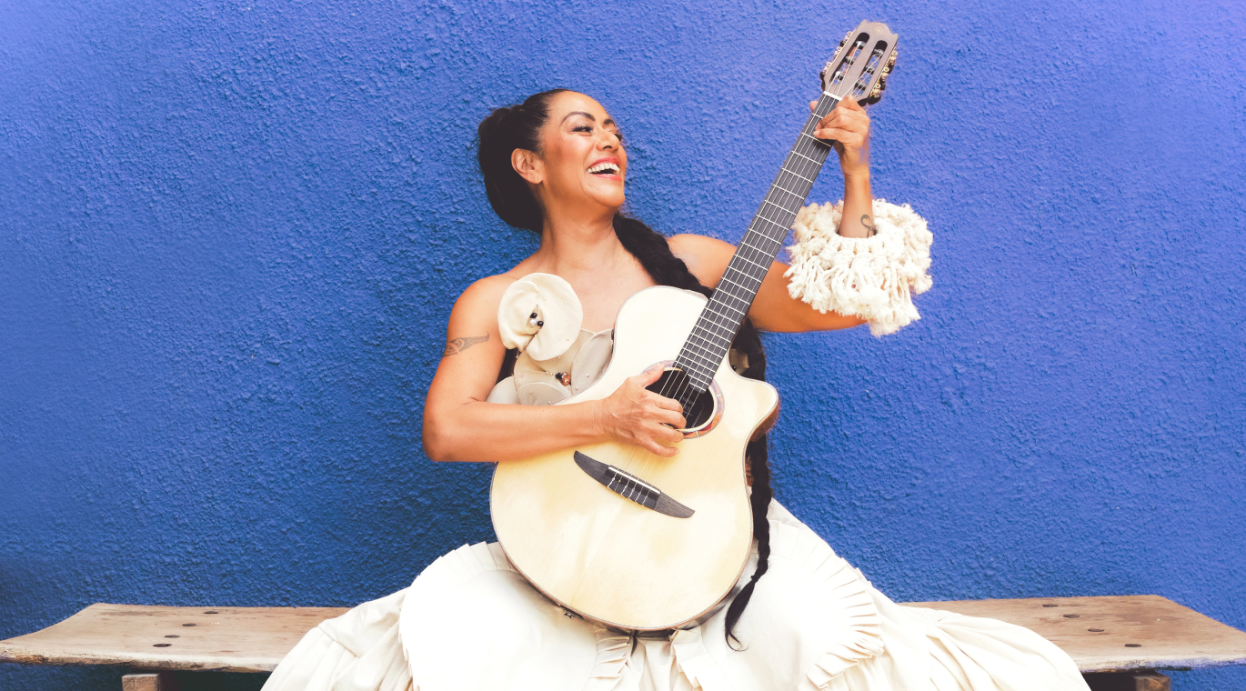 Mujer sonriente toca guitarra sentada en banca, con vestido blanco frente a pared azul intensa