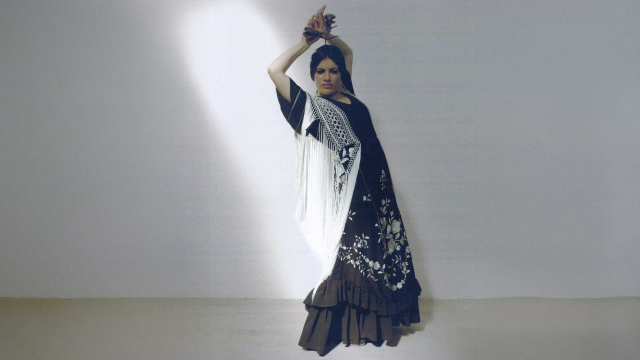Bailaora de flamenco con vestido negro bordado y mantón blanco de flecos, posando con los brazos alzados sobre fondo claro.