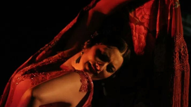Bailaora de flamenco iluminada con luz roja, inclinada hacia atrás bajo un mantón de flecos durante una actuación sobre fondo oscuro.