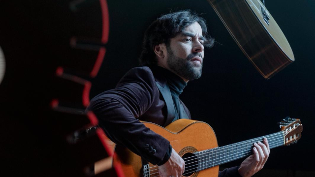 Guitarrista flamenco sentado en un escenario tocando una guitarra española, bajo iluminación teatral