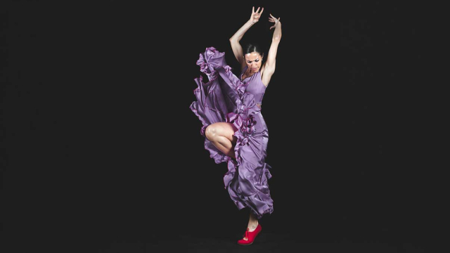 Bailaora de flamenco con vestido morado de volantes y zapatos rojos, ejecutando una pose dinámica sobre fondo negro
